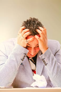 Businessman is suffering from a strong headache and feeling stressed while sitting at his desk with a crumpled piece of paper in front of him
