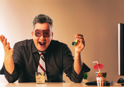 Businessman wearing a party mask drinking alcohol alone at his office desk trying to have fun issues of alcoholism in the workplace