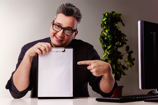 Professional is sitting at his desk, smiling and pointing at a blank clipboard. The image evokes concepts such as marketing, advertising, and business
