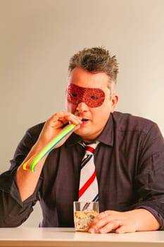 Businessman celebrates with a colorful mask and party blower, enjoying whiskey at the office in a festive costume, having a great time alone