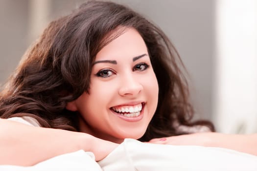 Young woman with long brown hair is smiling while relaxing on a white sofa in her living room, enjoying a peaceful moment at home