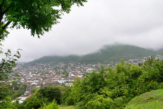 Shaki City in Azerbaijan sits nestled amidst rolling hills and abundant greenery, with clouds enveloping the landscape, creating a serene atmosphere.