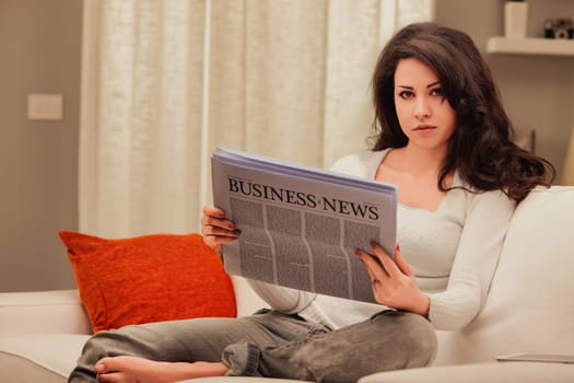Young woman reads a newspaper on her couch, looking serious. The cozy living room has warm natural light. She stays informed on finance and lifestyle