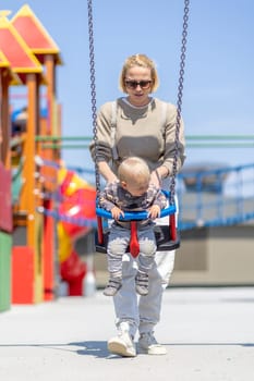 Mother pushing her infant baby boy child on a swing on playground outdoors