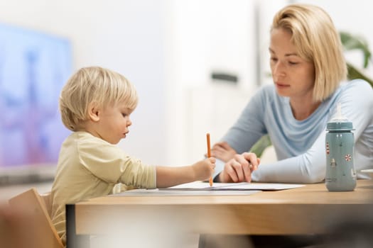 Caring young Caucasian mother and small son drawing painting in notebook at home together. Loving mom or nanny having fun learning and playing with her little 1,5 year old infant baby boy child