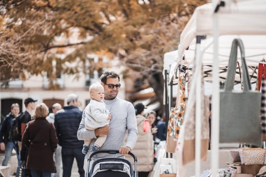 Father walking carrying his infant baby boy child and pushing stroller in crowd of people visiting Sunday flea market in Malaga, Spain