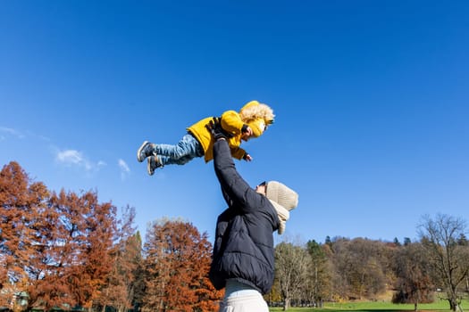 More, more,...mum, that's fun. Happy young mother throws her cute little baby boy up in the air. Mother's Day, Mather and her son baby boy playing and hugging outdoors in nature in fall