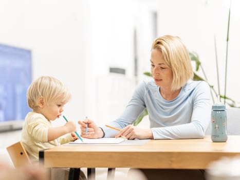 Caring young Caucasian mother and small son drawing painting in notebook at home together. Loving mom or nanny having fun learning and playing with her little 1,5 year old infant baby boy child