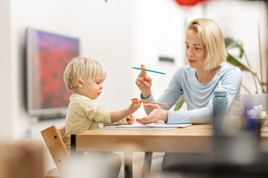 Caring young Caucasian mother and small son drawing painting in notebook at home together. Loving mom or nanny having fun learning and playing with her little 1,5 year old infant baby boy child