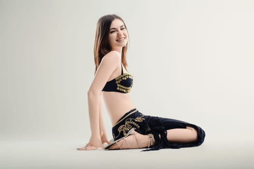 Young woman with long brown hair is kneeling on the floor, wearing a black and gold belly dancer outfit, and smiling. She is practicing a traditional exotic dance