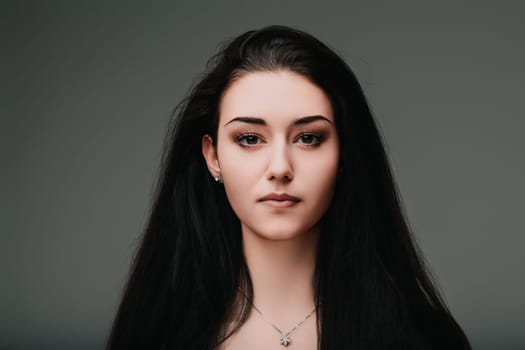 Young woman with long black hair is posing on a gray background, her serious facial expression suggesting deep thought and introspection