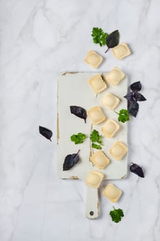 Overhead view of raw uncooked ravioli on a chopping board with scattered leaves of basil and cilantro