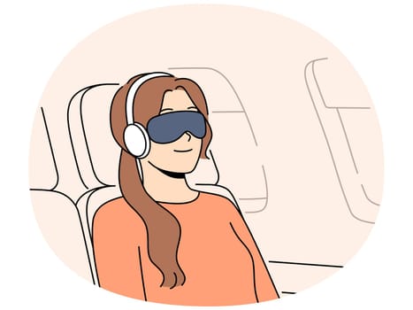 Woman passenger of flight with blindfold and earphones sleeps in airplane chair sitting outside porthole. Young girl first class passenger flies on business trip on airplane and smiles