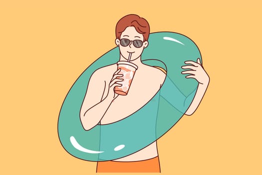 Man goes to beach and drinks cold cocktail, holding inflatable swimming ring in hand. Tourist guy enjoys relaxing near sunny pool at hotel and opportunity to sunbathe or swimming in warm sea