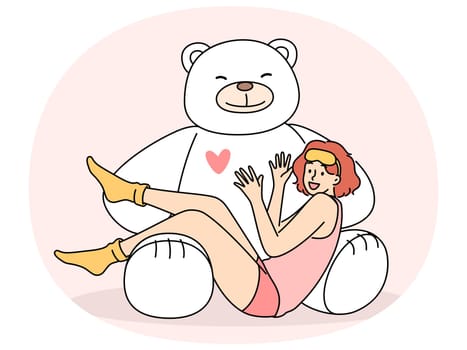 Giant teddy bear near sitting teenage girl in pajamas and sleep mask. Funny schoolgirl teen age looks at screen lying on feet of toy bear rejoicing at cute gift from parents or boyfriend
