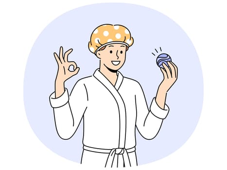 Man with bath bomb is dressed in bathrobe and bandage to protect hair from moisture. Young guy shows OK gesture recommending use of aromatic bath bomb while performing hygiene procedures.