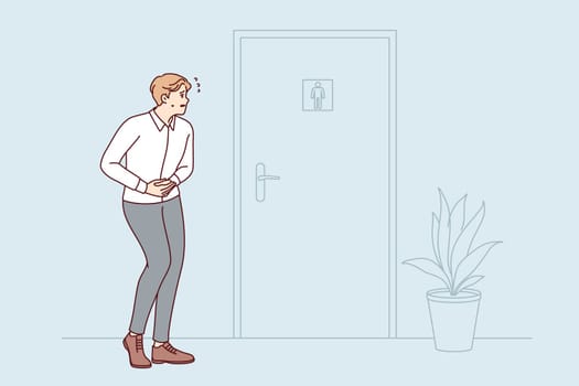 Man experiencing diarrhea stands near door to toilet room, holding stomach, feeling discomfort. Concept of queue at ws in office building due to lack of sufficient number of toilet stalls