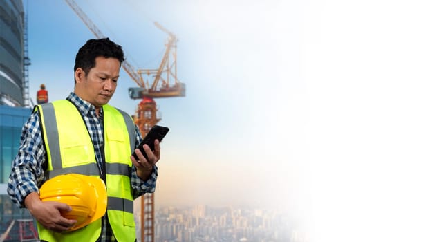 A man wearing a yellow vest and a hard hat is looking at a tablet. The tablet is displaying a construction site with cranes and buildings. The man is focused on the smartphone.
