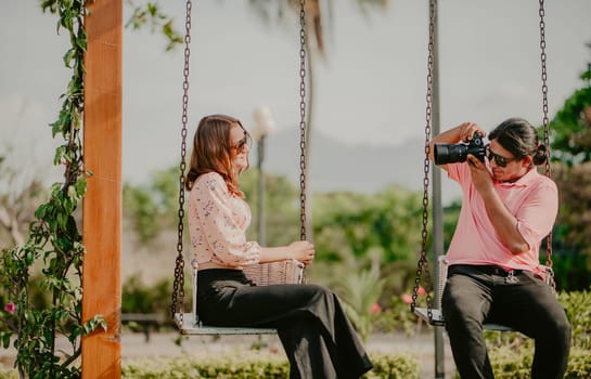 Boyfriend taking a photo of his girlfriend on a swing in a park. Boyfriend photographing girlfriend sitting on a swing
