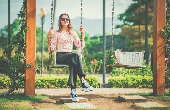 Cheerful girl in glasses relaxing on a swing in a natural park. Happy young woman sitting on a swing in a nature park