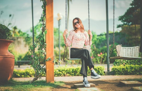 Happy young woman sitting on a swing in a nature park. Portrait of a smiling girl sitting on a swing in a nature park