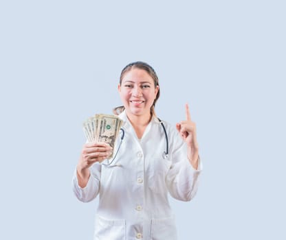 Smiling female doctor holding money and pointing up isolated. Happy young female doctor showing money and pointing at promo up