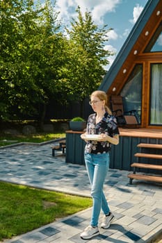A young woman carries plates to the patio table to lay out grilled food on backyard