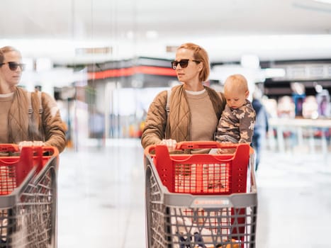 Mother shopping with his kid in the mall. Mother pushing shopping cart while holding and carrying her toddler baby boy child along mall corridor.