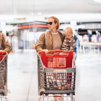 Mother shopping with his kid in the mall. Mother pushing shopping cart while holding and carrying her toddler baby boy child along mall corridor.