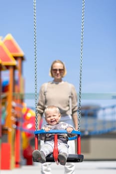 Mother pushing her infant baby boy child on a swing on playground outdoors