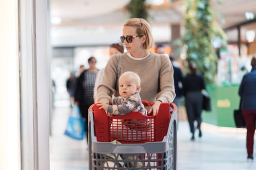 Mother shopping with his kid in the mall. Mother pushing shopping cart with her toddler baby boy child sitting in it along mall corridor.
