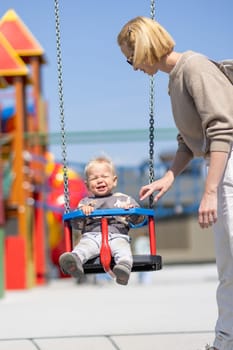 Mother pushing her infant baby boy child on a swing on playground outdoors