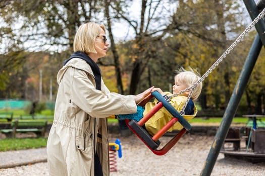 Mother pushing her infant baby boy child wearing yellow rain boots and cape on swing on playground outdoors on cold rainy overcast autumn day in Ljubljana, Slovenia.