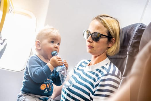 Mom and child flying by plane. Mother holding and playing with her infant baby boy child in her lap during economy comercial flight. Concept photo of air travel with baby. Real people
