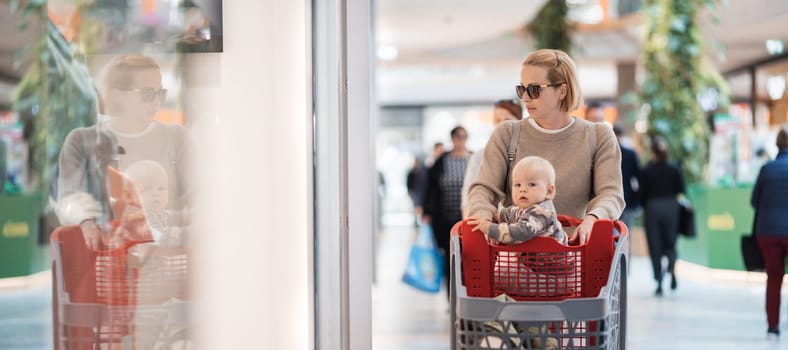 Mother shopping with his kid in the mall. Mother pushing shopping cart with her toddler baby boy child sitting in it along mall corridor.