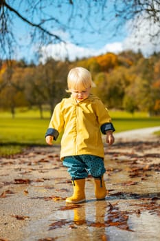 Sun always shines after the rain. Small bond infant boy wearing yellow rubber boots and yellow waterproof raincoat walking in puddles in city park on sunny rainy day