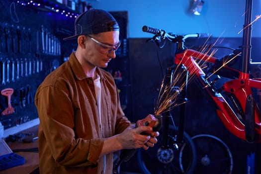 Young handsome man bike mechanic using circular saw for repair work. Closeup cropped shot on male hand sawing metal spare part in flow of sparks from angle grinder machine