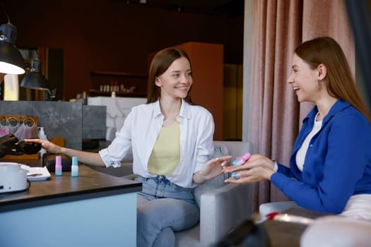 Young woman advising her friend trendy color of nail polish while getting manicure in spa salon. Feminine procedure and professional service concept