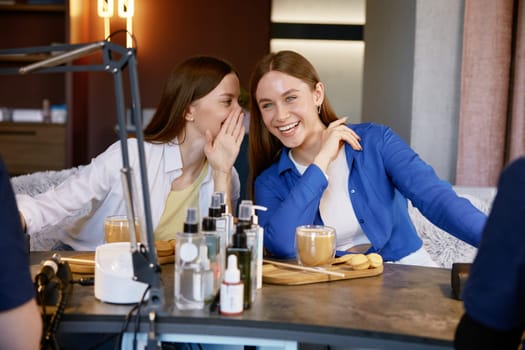 Young woman friends gossiping whispering secrets at nail studio. Two happy girls getting manicure spending fun time together at beauty salon