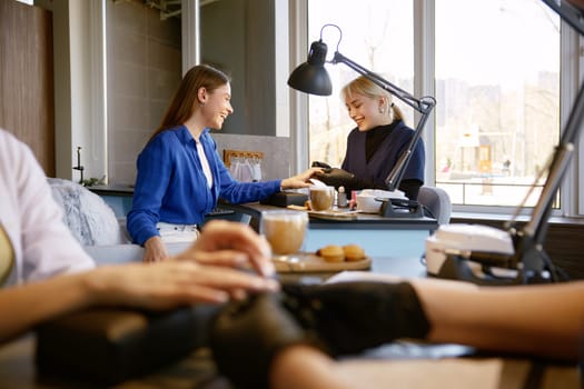 Female client getting high quality service at nail studio. Young woman customer drinking cappuccino eating sweet snacks while manicurist doing manicure