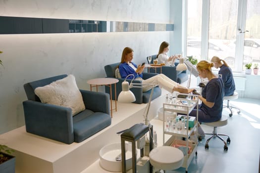 Elegant women clients relaxing with phones at luxury nail studio during high quality pedicure procedure