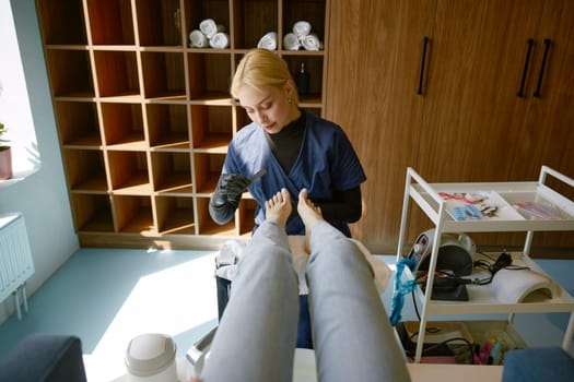 Friendly smiling pedicure specialist in uniform at work in modern beauty salon. Pretty female beautician providing foot health care procedure for client