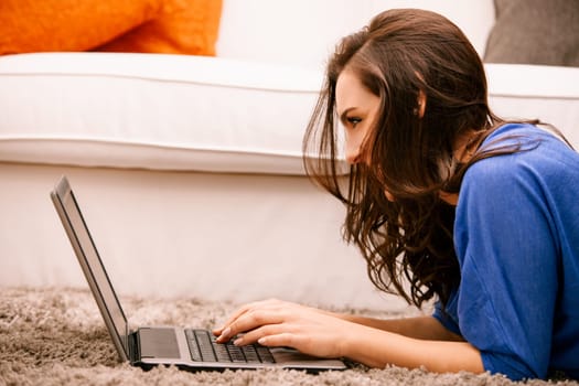 Young woman is lying on her stomach on the carpet and typing on her laptop, she is smiling and seems to be having fun