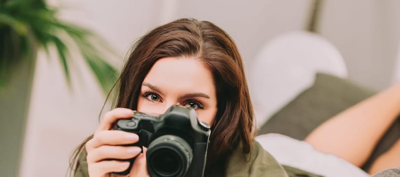 While on vacation, a young woman with brown hair and bright eyes playfully peers over her camera. The light-hearted moment captures her enjoyment of photography and leisure time