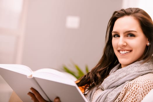 Young woman happily reading a book on a cozy sofa at home, surrounded by warmth and natural light, exuding relaxation and contentment