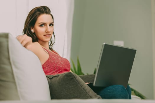 woman with a cheerful smile sits on a sofa, using a laptop. Her pink lace top and jeans complement the warm, inviting atmosphere created by her relaxed posture and the cozy environment