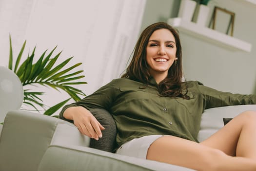smiling woman with long dark hair sits comfortably on a light-colored sofa. She wears an olive green shirt and white shorts, her relaxed posture reflecting a sense of ease and contentment