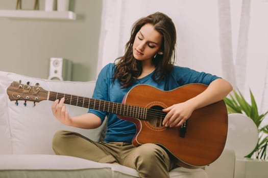 woman with long brown hair sits on a sofa, focused intently as she plays an acoustic guitar. Wearing a blue top and khaki pants, she exudes calm concentration in a cozy, serene environment