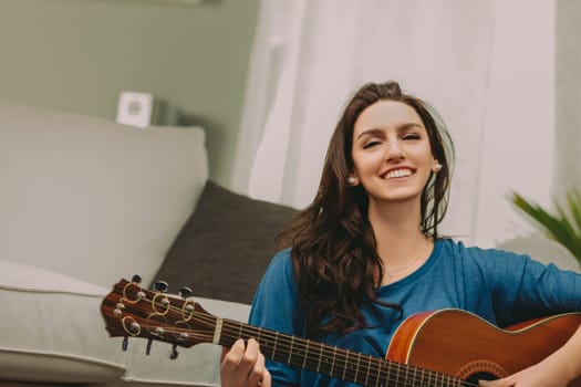 Smiling woman, wearing a blue shirt and khaki pants, sits comfortably on a sofa, concentrating on playing an acoustic guitar. The calm surroundings enhance her serene and focused demeanor
