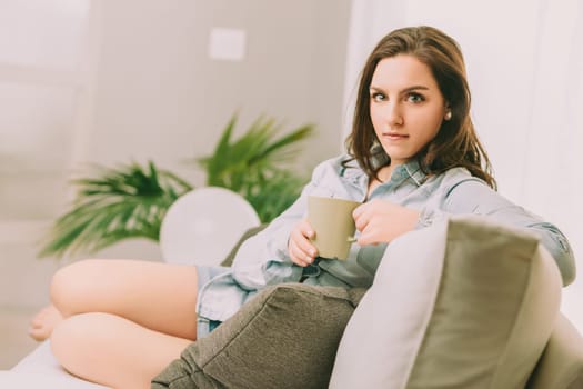 Sitting on a comfortable sofa, a woman with a pensive look holds a green mug. Her relaxed posture, denim shirt, and the surrounding greenery create a tranquil and contemplative atmosphere